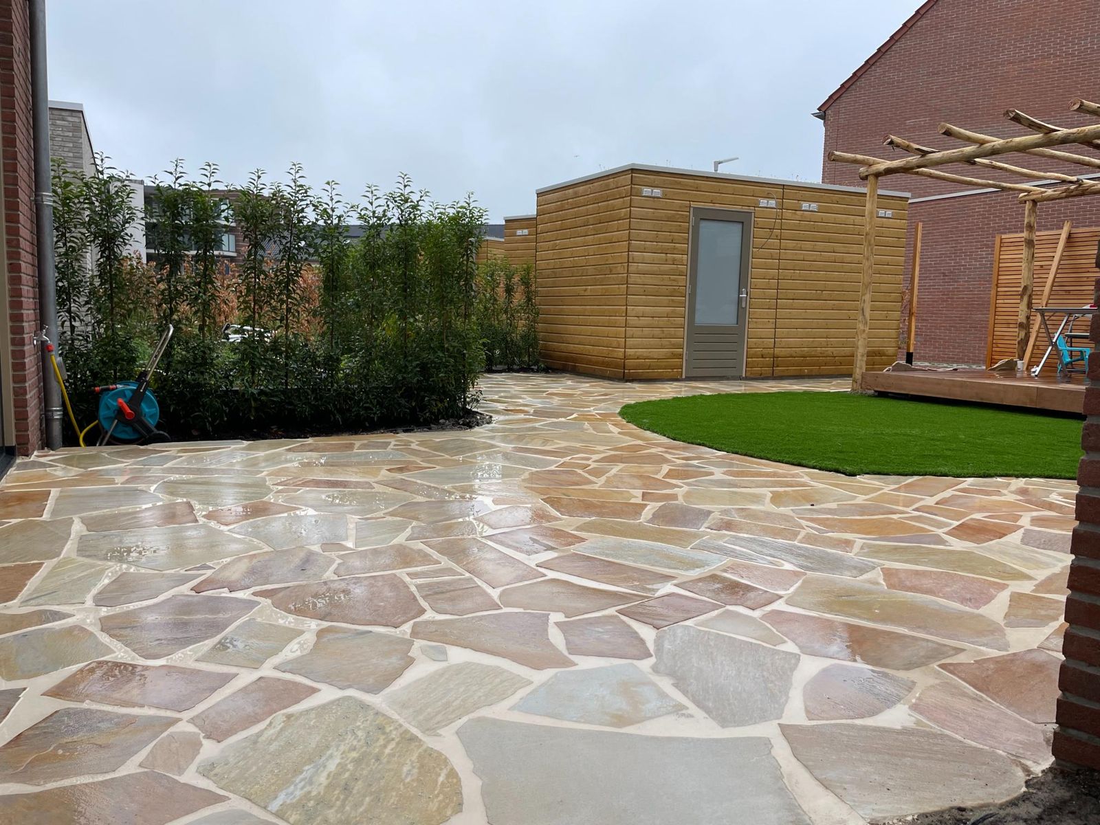 Achtertuin met een pad en terras van onregelmatige natuurstenen platen, kunstgras en een houten tuinhuis