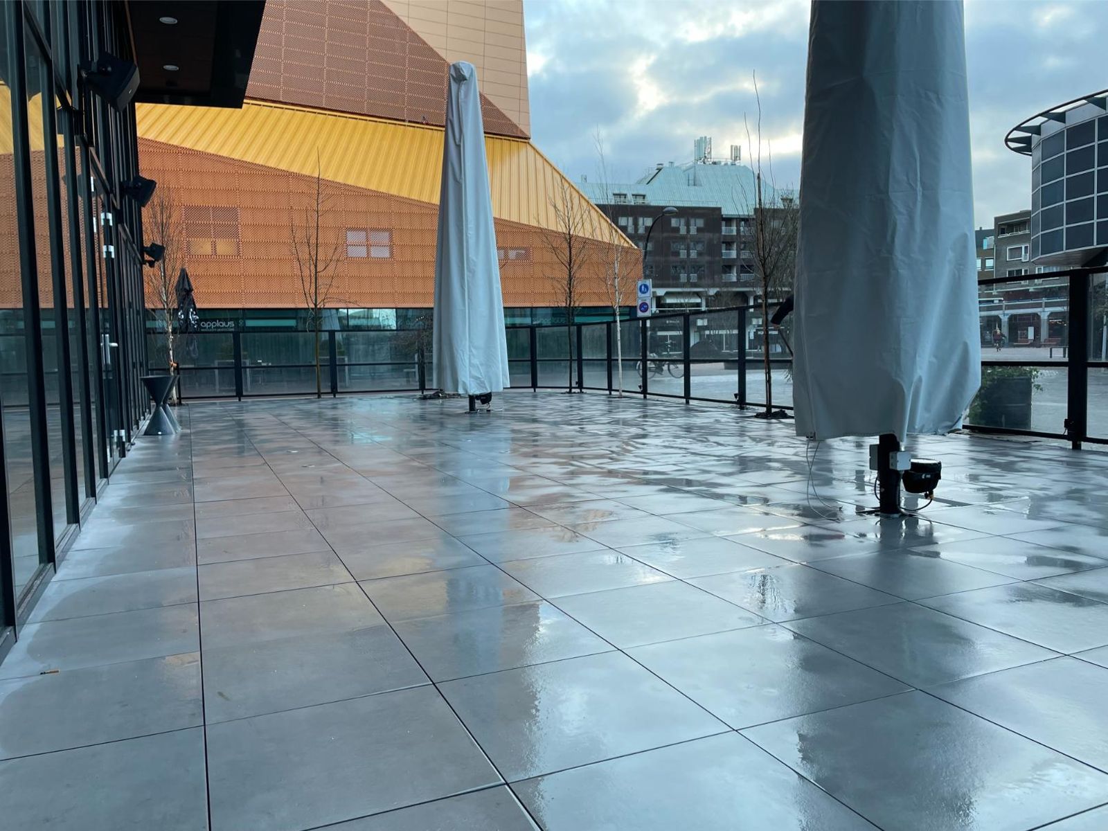 Ruim terras van grote grijze tegels met glazen balustrade en parasols, nat na een regenbui
