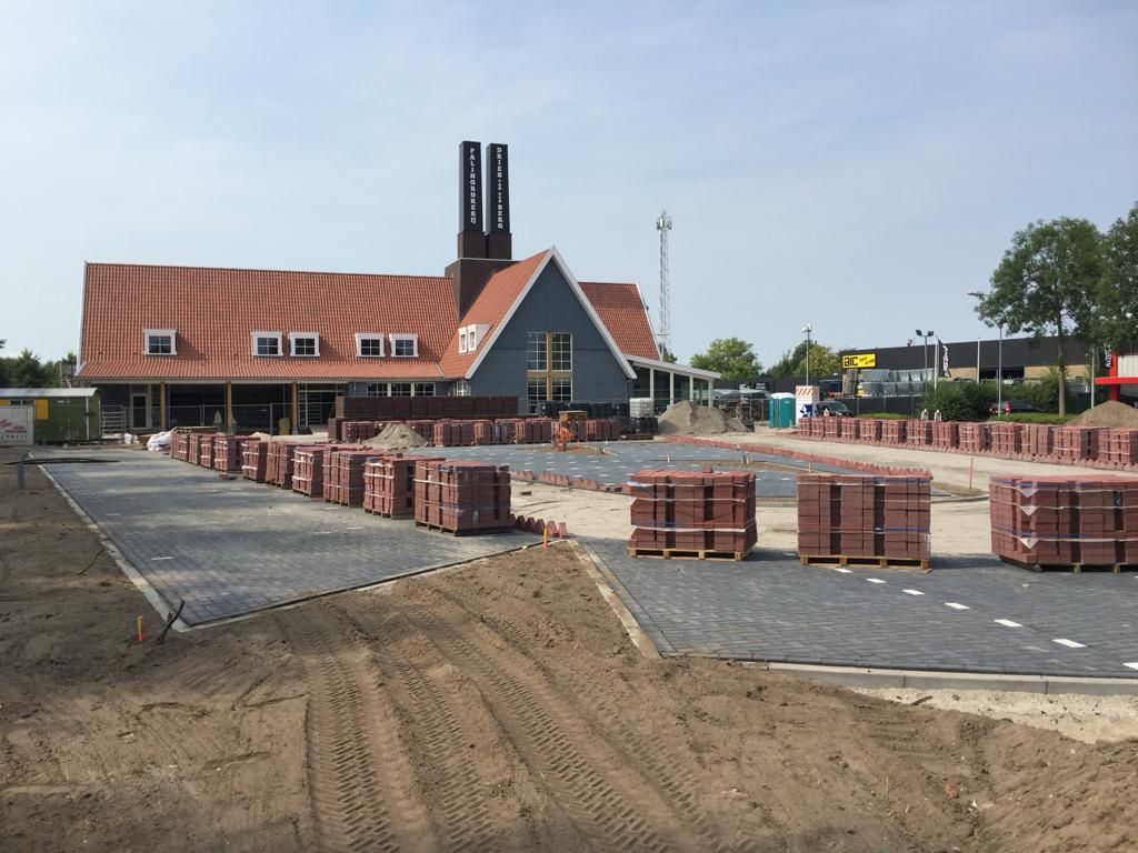 Groot terrein in aanleg met deels gelegde grijze bestrating, pallets met stenen en zand op de voorgrond