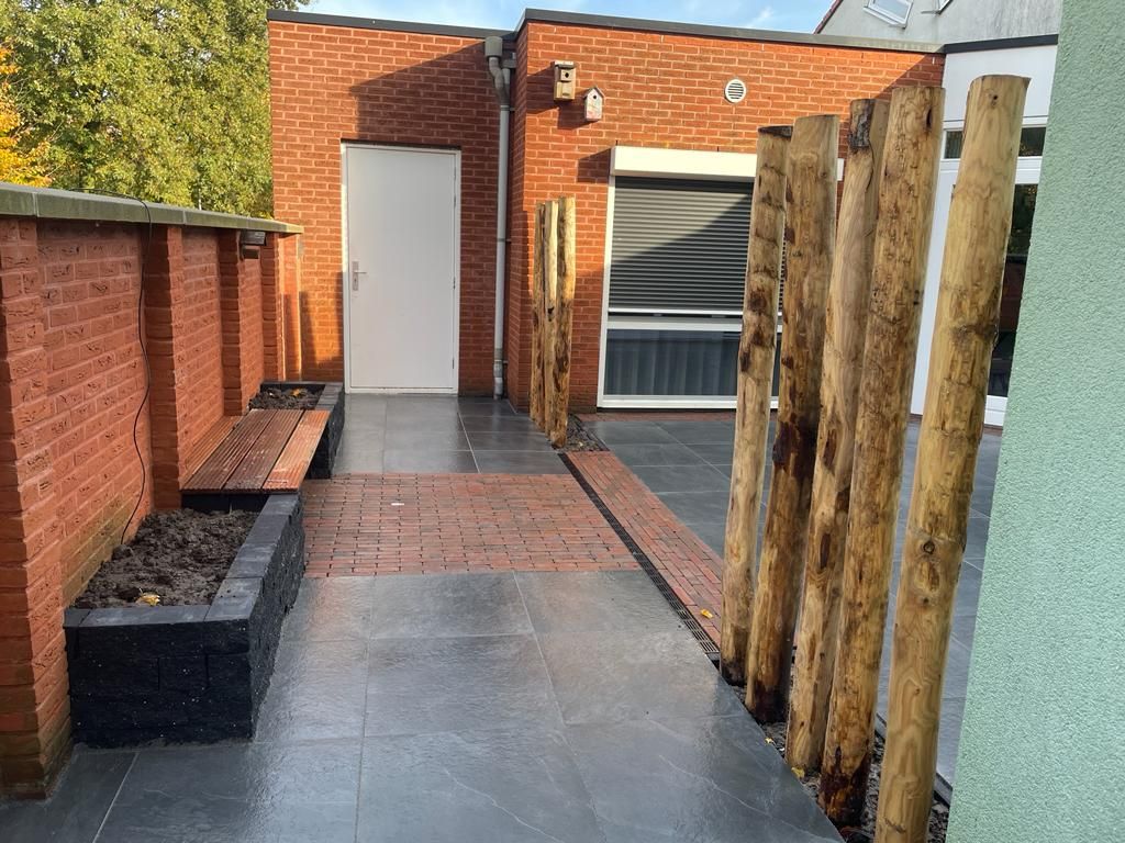 Smal zijpad van grote donkergrijze tegels met een rand van rode klinkers langs een bakstenen muur en houten palen