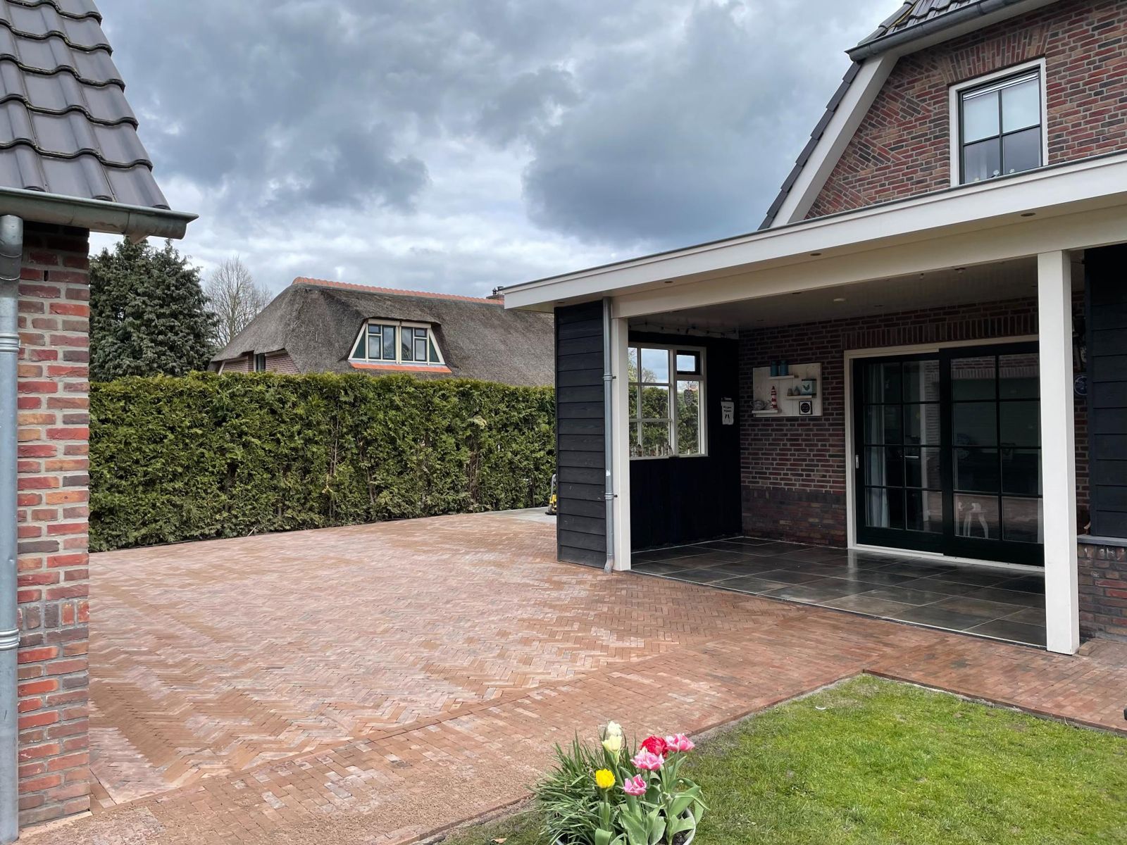 Terras van rode gebakken klinkers voor een donkere overkapping met glazen schuifpui