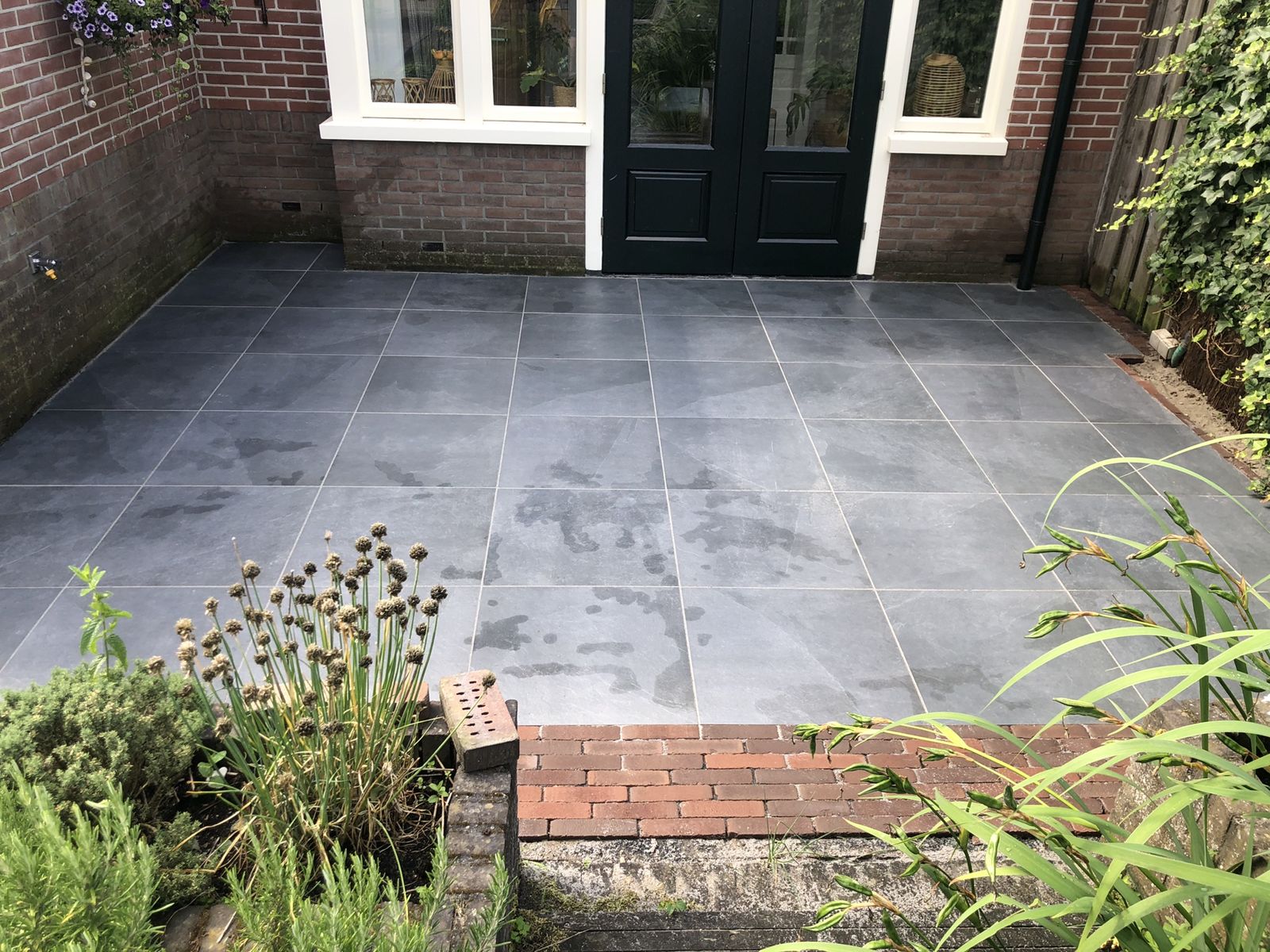 Terras van grote antracietkleurige tegels voor openslaande deuren, met een rand van rode klinkers en beplanting